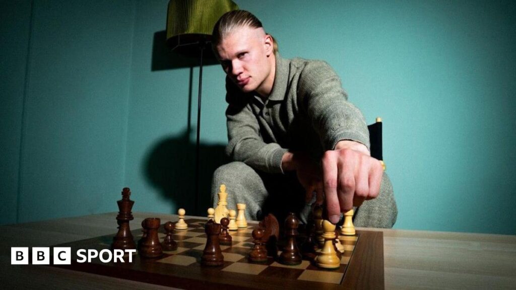 Erling Haaland looks into the camera as he moves a white bishop on a chess board.